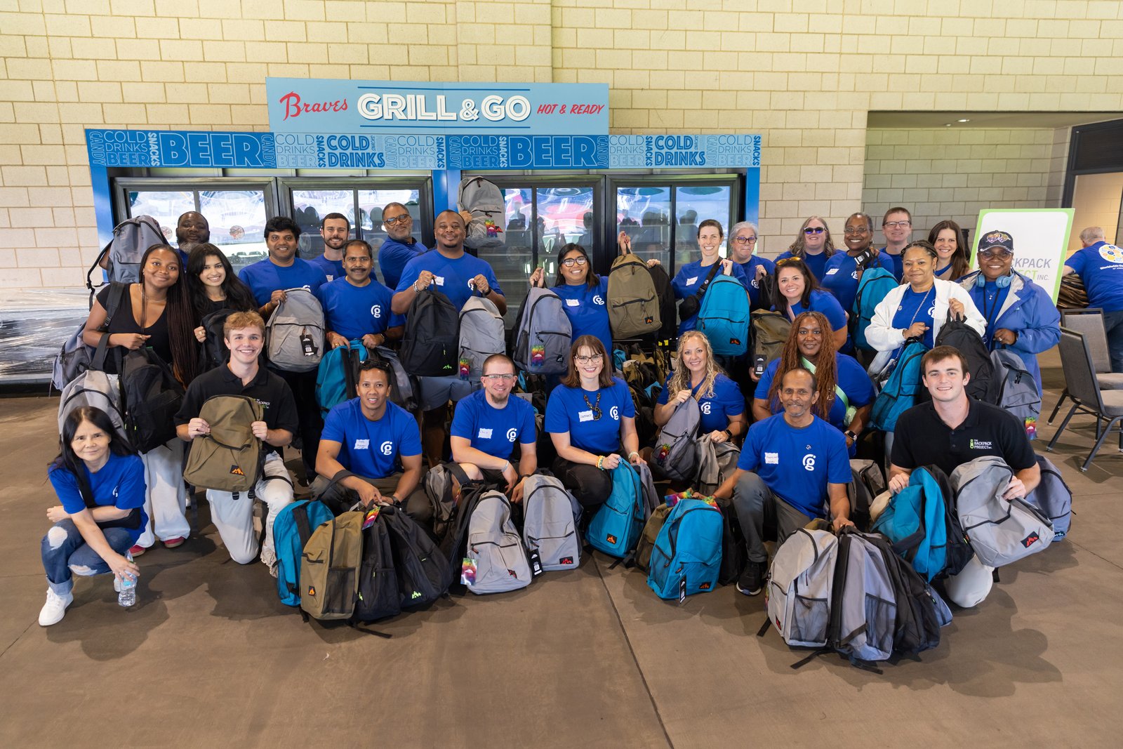 Volunteers in Georgia Power shirts and Backpack Project polos pose with packed backpacks at a Braves Grill & Go stand.