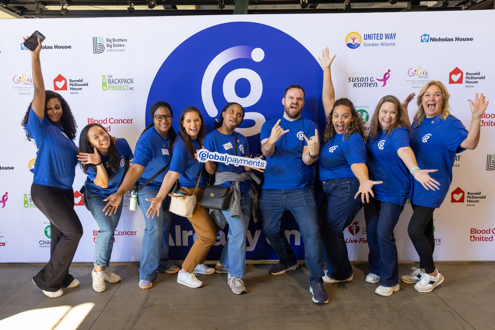Volunteers pose in front of a partner step-and-repeat that includes The Backpack Project Inc. logo.