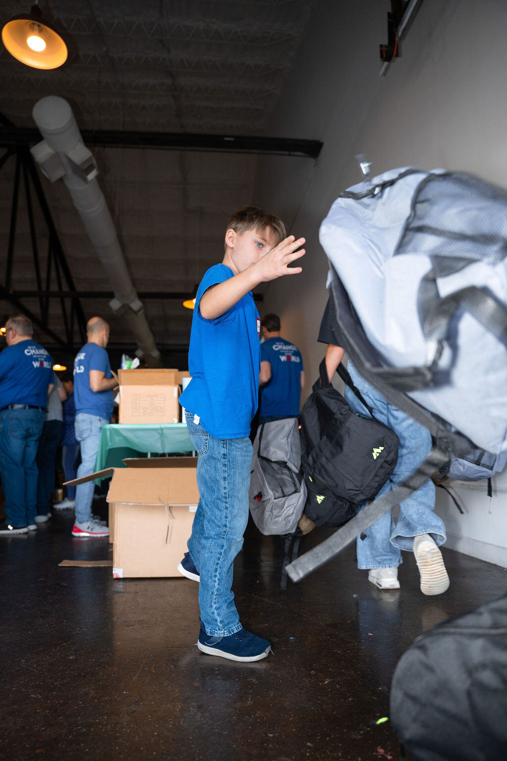 Volunteers in matching shirts pack backpacks at long tables.