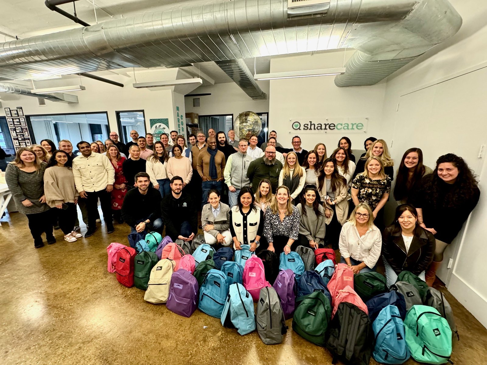 A large volunteer group poses behind rows of packed backpacks at Sharecare.