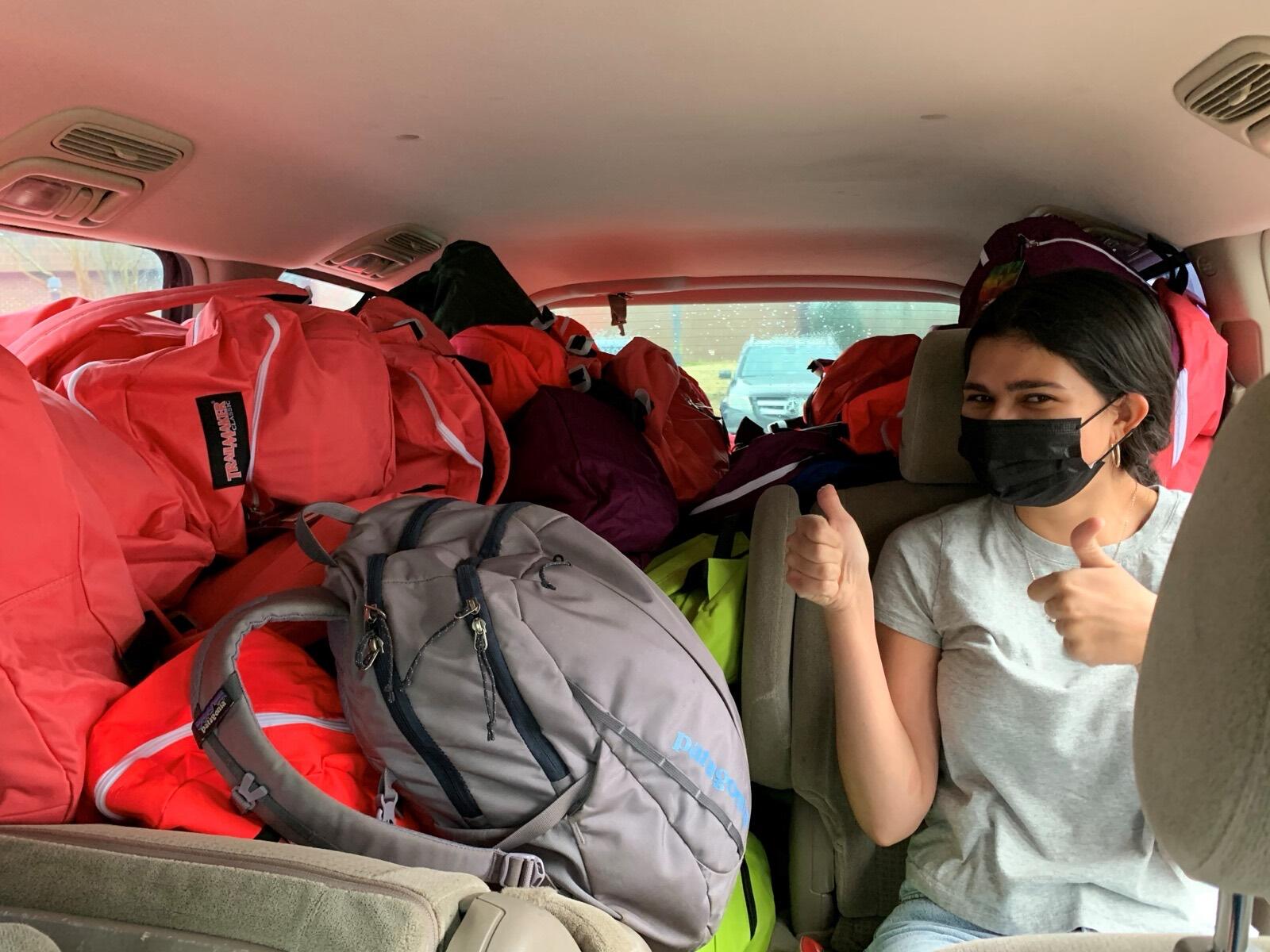 A volunteer in a car full of packed backpacks gives a thumbs-up before a delivery.